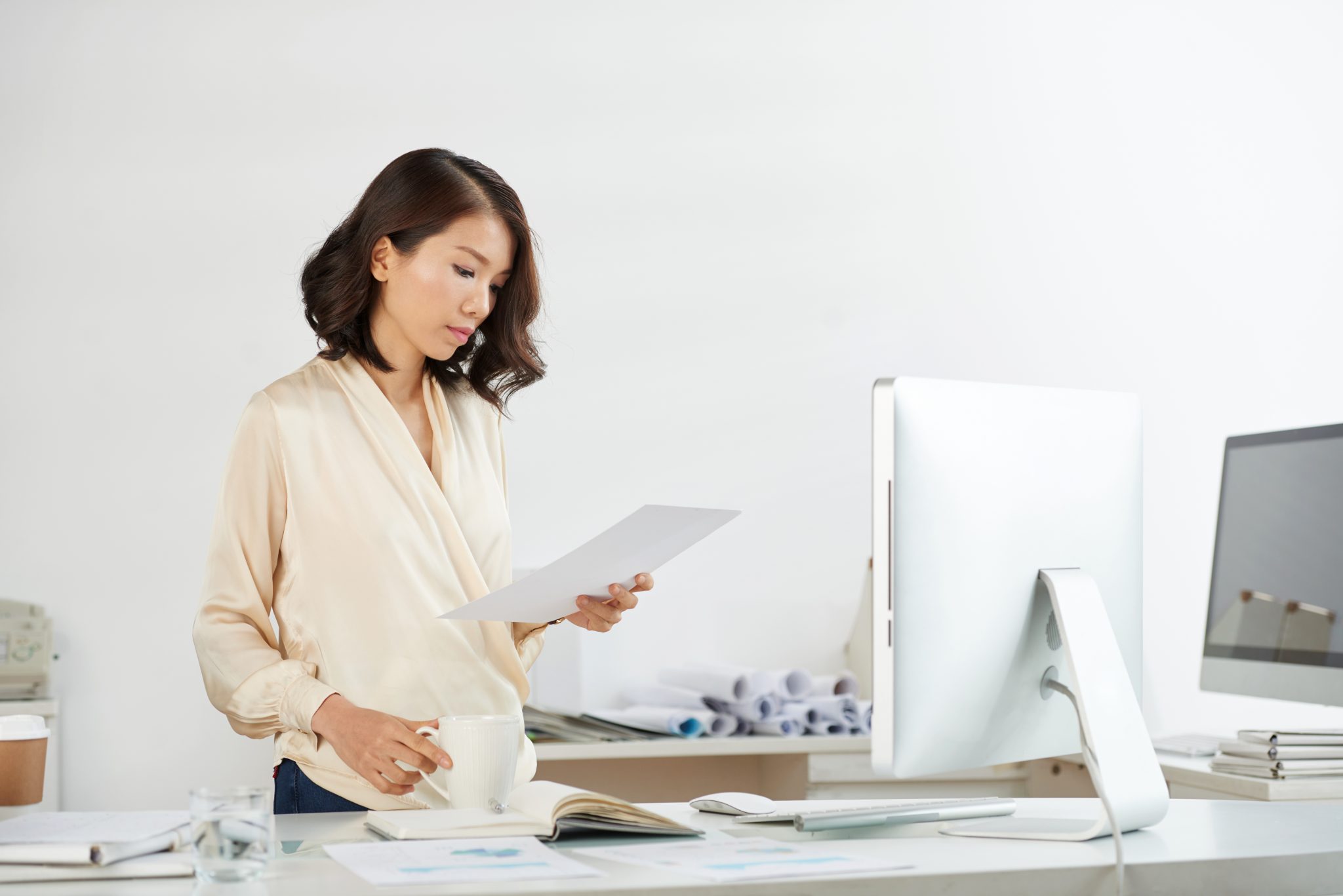 a female employee is on her feet, proofreading a document in front of her station with a cup of tea on her other arm.
